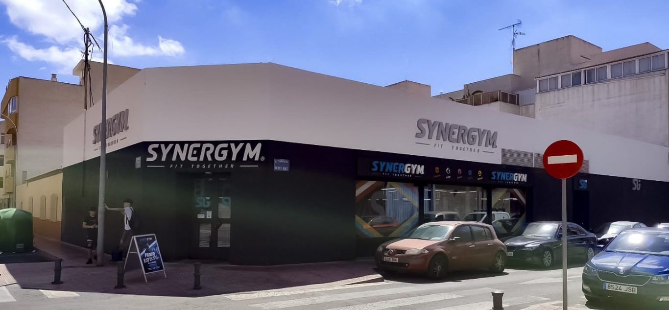 Edificio de gimnasio con fachada moderna y un cartel SYBERGYM grande en la entrada.