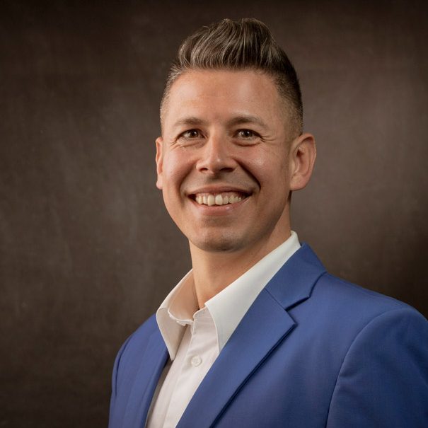Hombre sonriente con cabello corto, vestido con chaqueta azul y camisa blanca.
