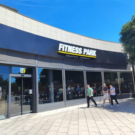 Exterior del gimnasio "Fitness Park" con personas entrando y saliendo bajo un cielo azul.